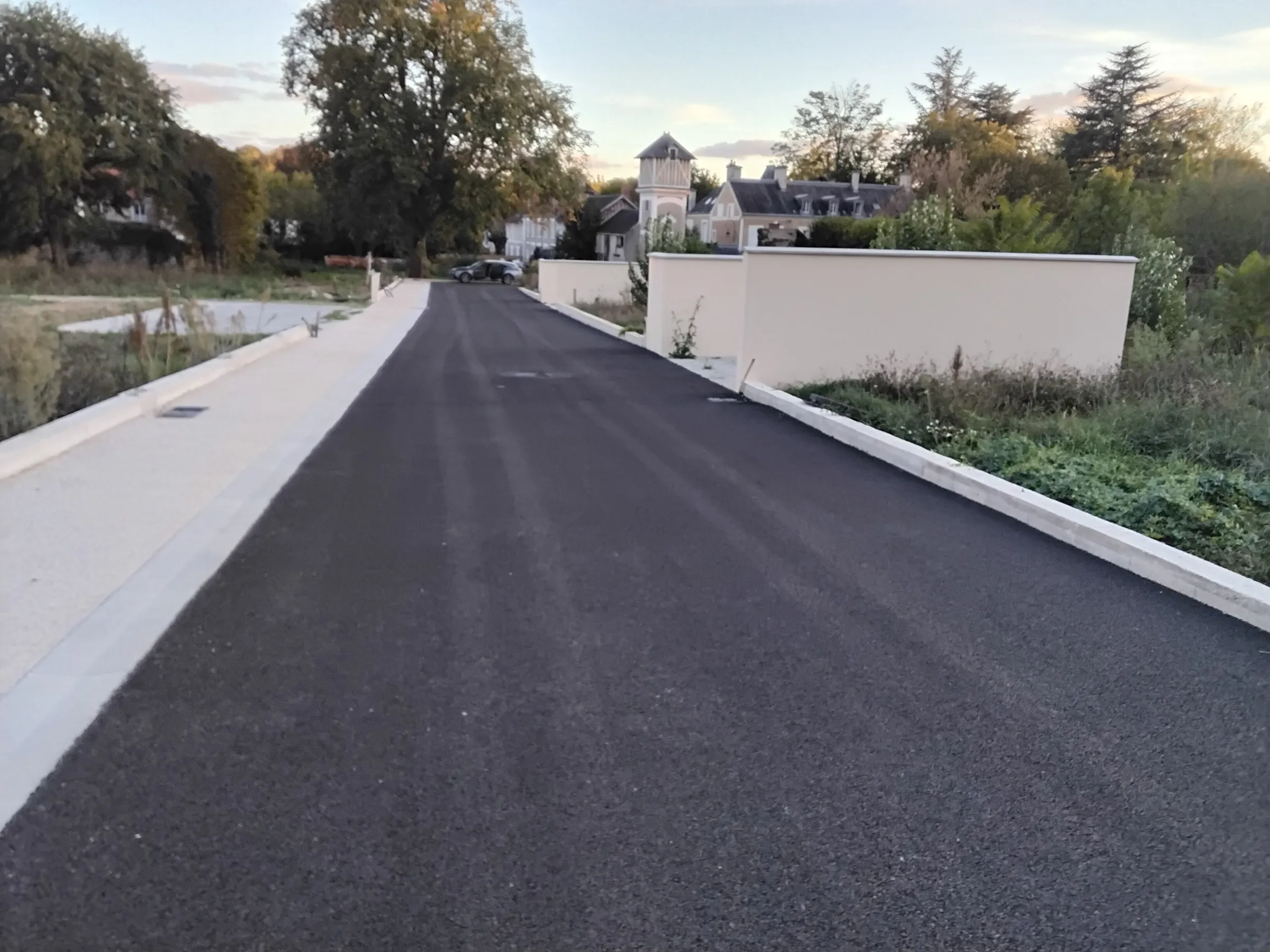Boissettes : la rue du lotissement terminée, enrobé neuf et trottoir, maisons du village en fond
